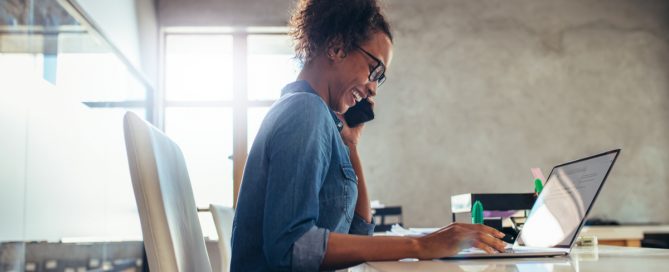 woman at desk on laptop using phone